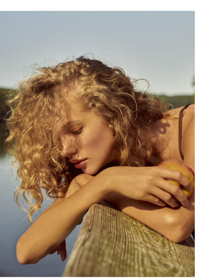 Portrait in golden light — curls, calm, an apple held against a wooden ledge by the water.